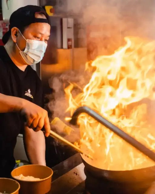 Chef en pleine préparation d’un wok thaï enflammé dans la cuisine de Bangcook, avec des flammes spectaculaires et des plats en cours de cuisson