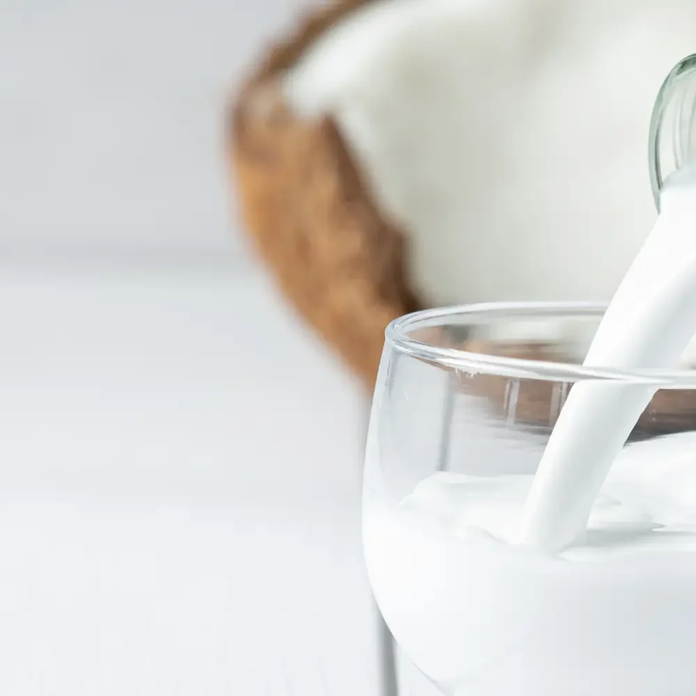 Noix de coco fraîche coupée en deux à côté d’un bol de lait de coco sur fond blanc lumineux avec texture bois naturelle. Ambiance minimaliste et tropicale